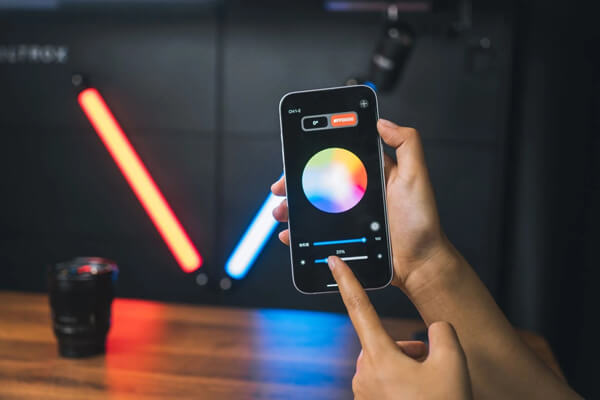 Рука держит смартфон с открытым приложением WeeylitePro, на экране видны цветовое колесо и ползунок яркости, на заднем плане размыто светятся две лампы Viltrox K60.